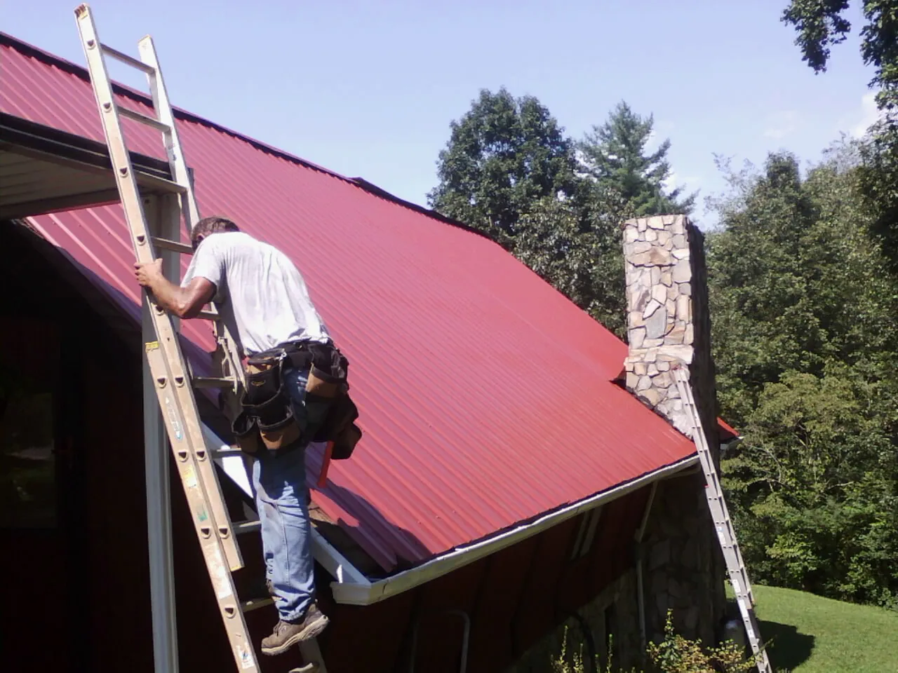 Roofer on a ladder beside a red metal roof and stone chimney — Soffit & Fascia Repair by Roofing Brothers in Eastview